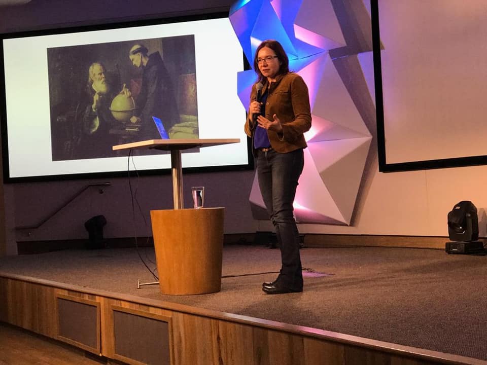 A woman stands at a podium orating in front of a power point presentation.