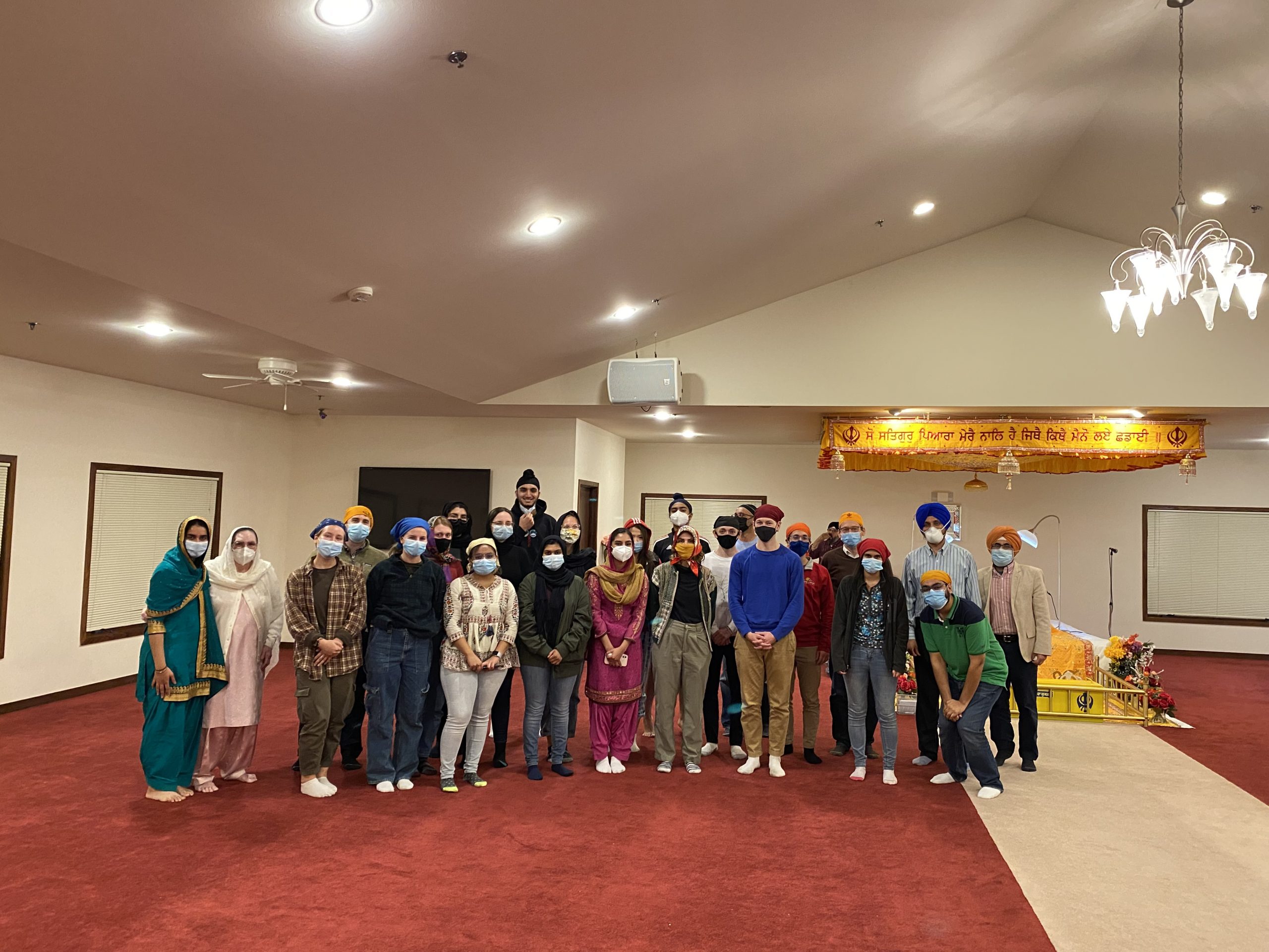 People wearing COVID masks and gathering inside a large room with red carpet and a hold sign.